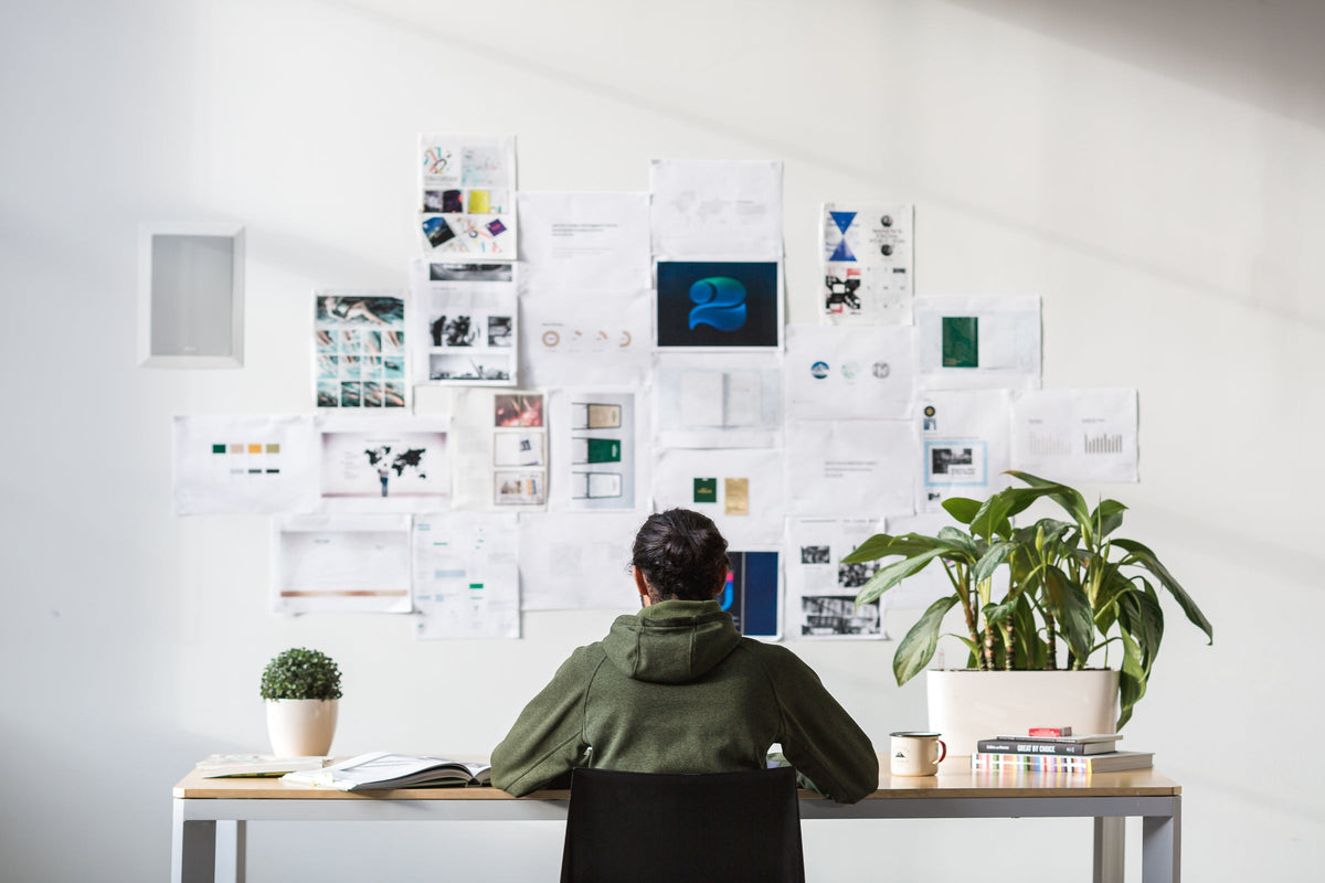 Person am Schreibtisch vor einer Wand mit Entwürfen und Moodboard